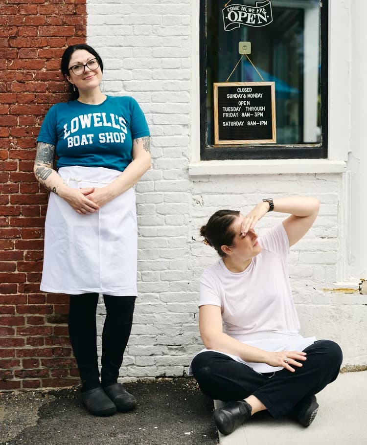 Emma and Melissa outside Middle Street Foods, standing and sitting on the sidewalk beside the shop's open sign